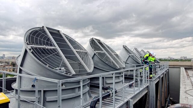 Workers fix large fans on top of a building in a city on a cloudy day