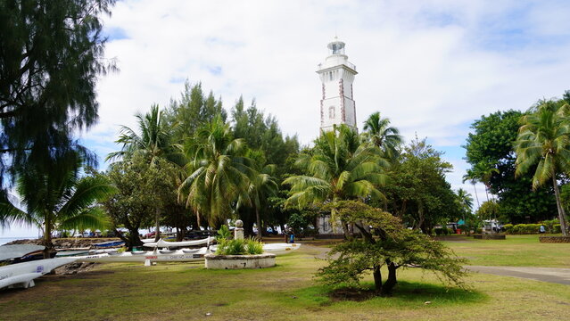Scenic view of the historic 1867 Pointe V&eacute;nus Lighthouse in Mahina, Tahiti, nestled within a lush green park filled with palm trees and outrigger canoes under a partly cloudy sky