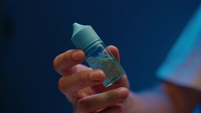 Close-up of hand holding and shaking small bottle of e liquid. Clear fluid mixes inside container. Moody blue lighting creating modern, technological, scientific atmosphere. Shooting in slow motion.
