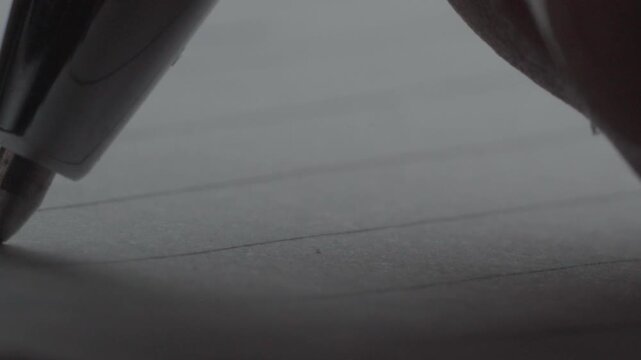 Close-up of a pen writing on a sheet of paper. The writing action is shown in detail, focusing on the pen tip, handwriting, ink marks, and paper surface
