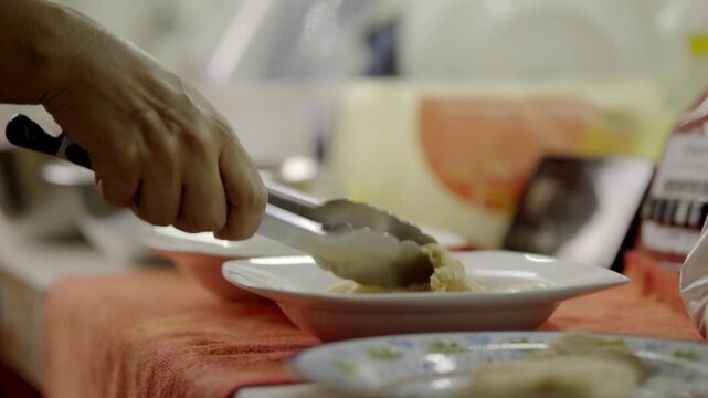 Close-up of a hand lifting a piece of cooked chicken with metal tongs over a white plate. The food is being prepared and served at a dining table indoors