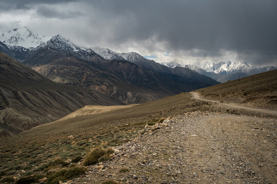 View of the Wakhan Corridor featuring a rugged dirt road winding through dramatic mountain ranges with snow-capped peaks under a cloudy sky in Badakhshan, Afghanistan.