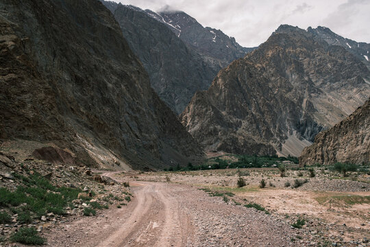 View of the rugged Bartang Valley landscape with a winding dirt road leading towards towering rocky mountains under a cloudy sky in Chah Ab, Gorno-Badakhshan Auto, Tajikistan.