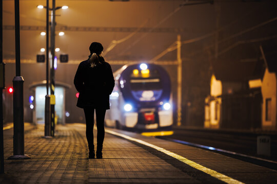 Woman looking at arriving train at empty railway station at night. City life and commute by public transportation
