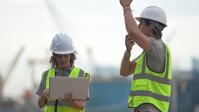 Port Workers Coordinating: Two construction workers in hard hats and safety vests are coordinating at a port with cranes in the background. One is holding a laptop.