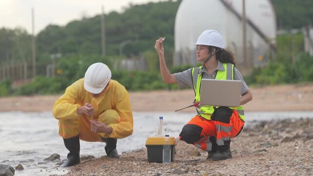 Environmental Sample Collection: Two scientists in protective gear and hard hats collect water samples from a shoreline, diligently recording data on a laptop.
