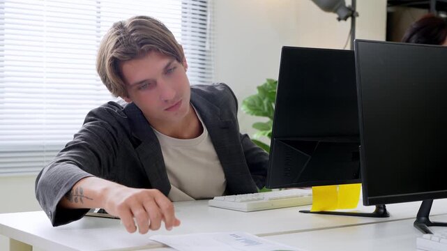 Exhausted Professional at Desk: A young man in a suit slumps over his desk, looking drained and fatigued while working late on his computer in a modern office environment.