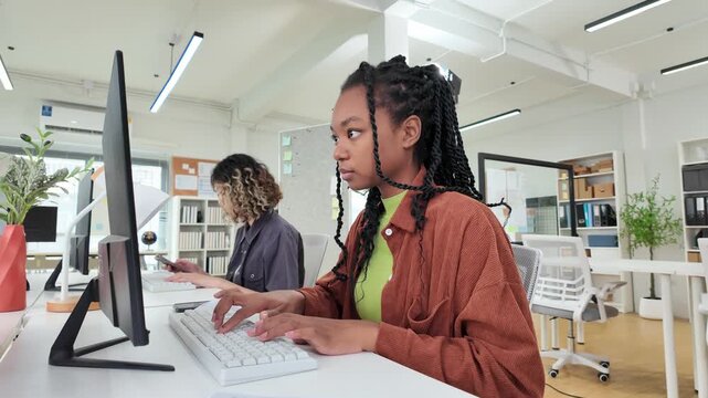 Focused Developer: A young woman with braided hair diligently works on a computer in a bright, modern office space, symbolizing dedication and technological advancement.
