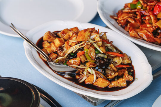 Chicken pieces with vegetables in dark savory sauce served on white oval plate. Asian style meal with spoon and colorful ingredients creates restaurant lunch and dinner food background.