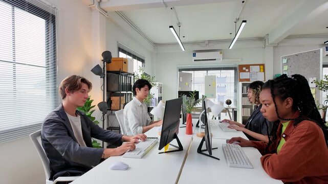 Diverse Team Collaborating in Modern Office: A vibrant mix of professionals, men and women from various backgrounds, are focused on their computer screens, working together in a brightly lit.