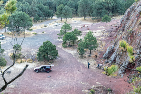 Vehicles parked in a red-earth clearing surrounded by pine forest in Cumbres del Ajusco National Park in Mexico City, Mexico, with visitors gathered near a rocky outcrop.