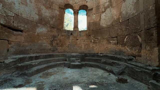 Slow upward camera tilt inside the ancient stone interior of Alahan Monastery in Mut, Mersin, T&uuml;rkiye, revealing curved stone walls, arched openings, a historic column and blue sky above. Early Christ