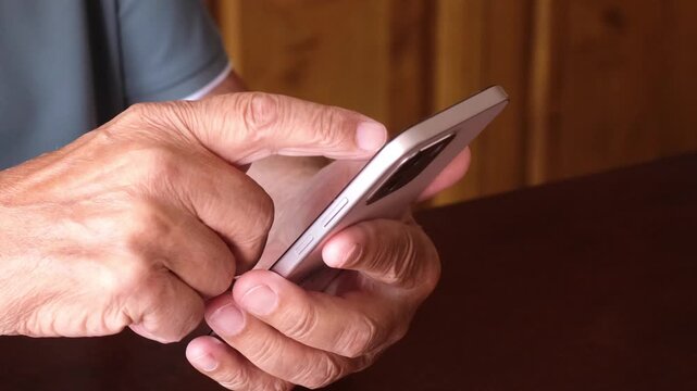 Close-up of an elderly man's hands holding and using a modern smartphone, swiping and tapping the touch screen while browsing online content or using an application at home