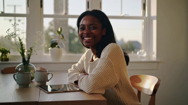 Smiling woman in chunky sweater at table camera starting leaning waving at lens, with tablet, mugs
