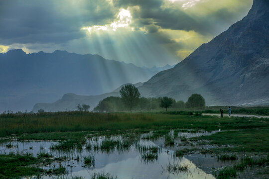 View of Shigar River with dramatic sun rays breaking through clouds over the rugged mountains, grassy wetlands, and trees in Shigar, Gilgit Baltistan, Pakistan.