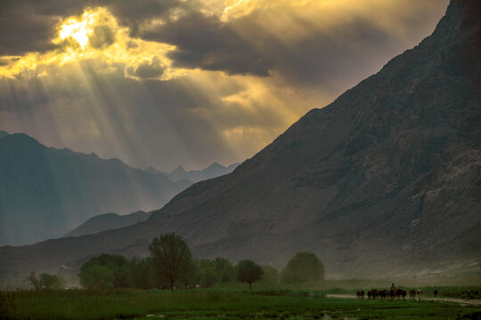 View of the Shigar River valley landscape with dramatic sunbeams breaking through clouds over the rugged mountains and lush green trees in the foreground in Shigar, Gilgit Baltistan, Pakistan.