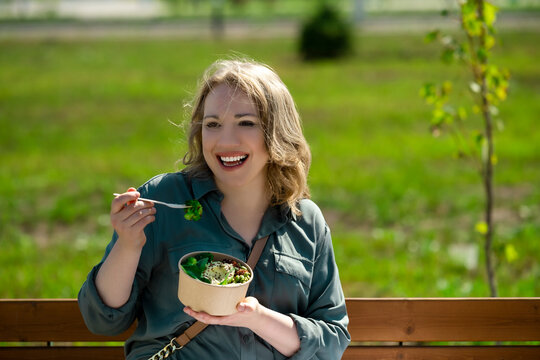 Happy woman eating healthy street food super bowl outside with wooden fork. Joyful plant based nutrition lifestyle. Smiling lady expressing positive emotions enjoying fresh green vegetable lunch.