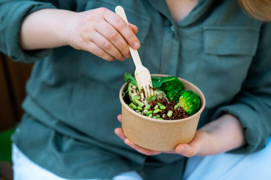 Female hands holding healthy street food super bowl with fresh green vegetables. Balanced plant based nutrition choice, vegan business lunch from eco paper container outside.