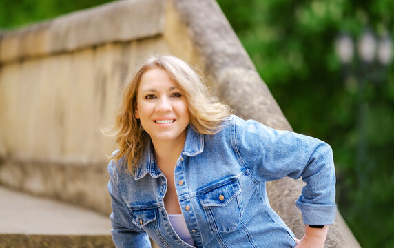 Adult blonde woman in denim jacket on stone stairs in park. Outdoor lifestyle concept for casual fashion, autumn portrait, and positive emotion.
