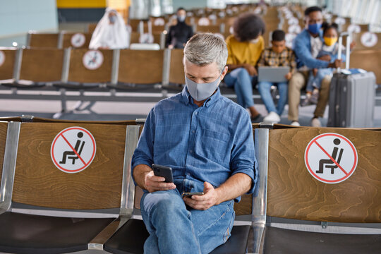 A man sits in an airport waiting area, looking at his phone, with social distancing signs on the seats around him