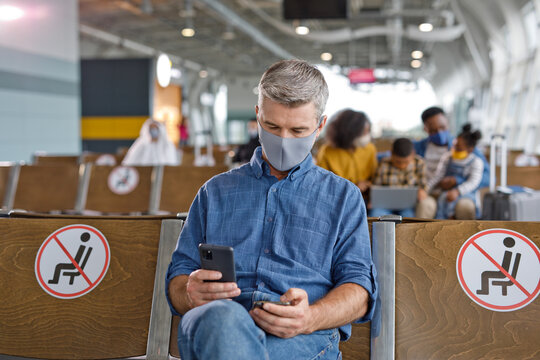 A man sits in an airport waiting area, looking at his phone, with social distancing signs visible on the seats