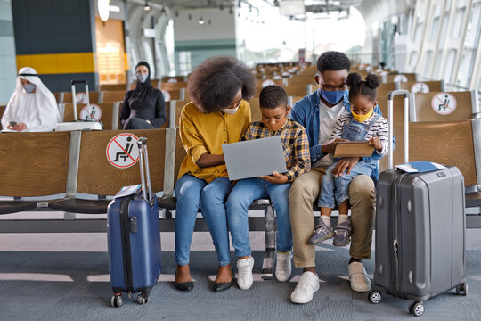 A Black family with two children sits in an airport terminal, wearing face masks and using electronic devices while waiting for their flight