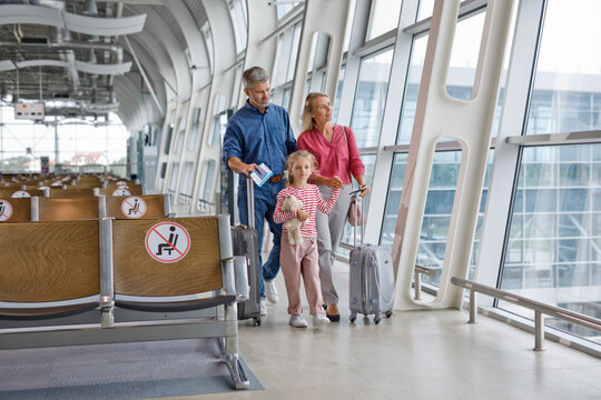 A family of three, parents and a young daughter, are walking together through a modern airport terminal with suitcases in tow
