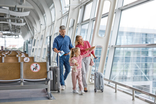 A mother, father, and daughter are walking through a bright airport terminal with their suitcases, gazing out the large windows