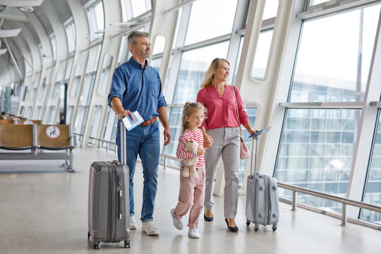 A family of three, parents and a young daughter, are walking together through a modern airport terminal holding their travel documents and suitcases