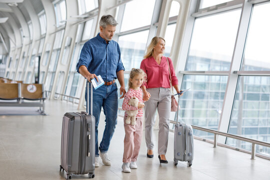 A family of three, parents and a young daughter, are walking together through a modern airport terminal holding their travel documents and suitcases