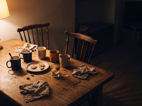 A kitchen table shows empty cups, a plate with crumbs, and cloths scattered after a gathering. The scene has dim light and simple wooden furniture that adds to the cozy atmosphere