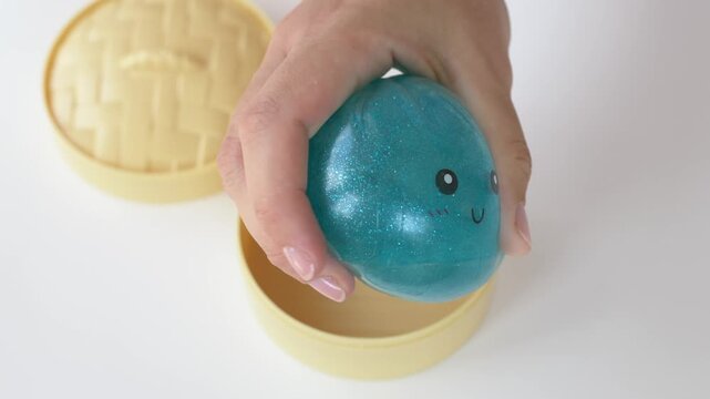 Hand lifts blue dumpling squishy toy from yellow container, revealing glossy texture and cute face. Product demonstration highlights tactile play, collectible appeal, colorful design, sensory