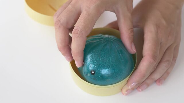 Hand presses turquoise squishy dumpling toy into round bamboo steamer box for demonstration in warm tone. Glossy character with cute smiling face shows soft texture fitting inside container