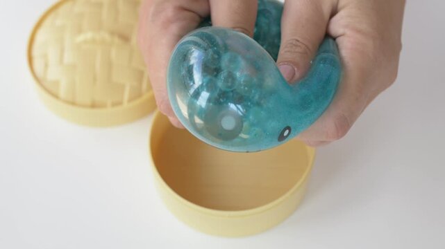 Hand lifts glossy turquoise squishy dumpling toy out of round bamboo steamer box quickly in home setting. Shiny wet look surface catches light while revealing cute smiling printed face closeup 