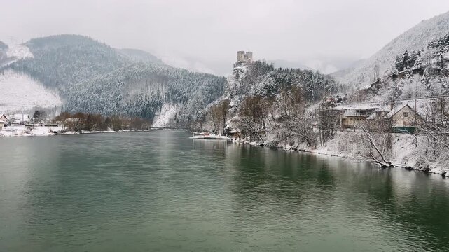 Medieval Strecno Castle on a snowy cliff above the Vah river in winter. Historic 4K landscape of a Slovakian fortress with an old river ferry and mountains in the Mala Fatra region, Slovakia.