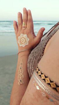 Bohemian woman adorned with a white mandala hand design, layered jewelry and patterned clothing holds a meditative gesture near waves, expressing mindfulness and calm coastal serenity
