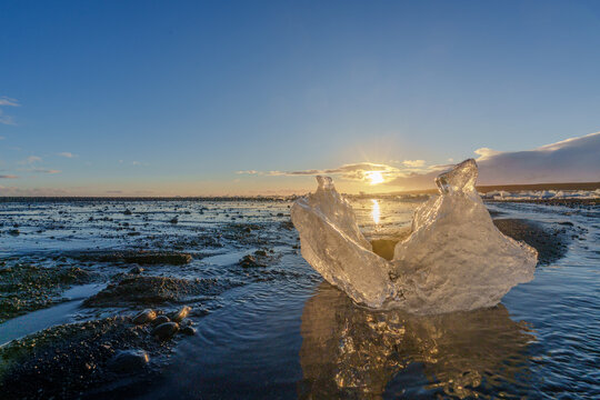 View of a translucent ice chunk resting on a dark volcanic sand beach at sunset with the sun reflecting on the wet shore in Iceland.