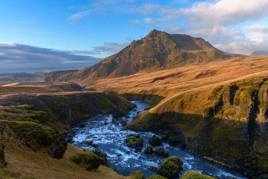 View of a river flowing through a rocky valley with golden grass and mossy boulders under a clear sky in Iceland.
