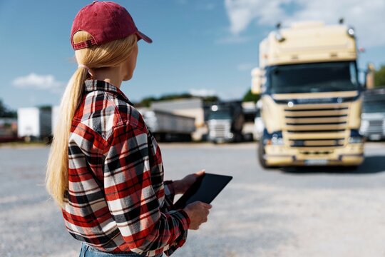 Woman truck driver in plaid shirt and cap holding tablet near semi-trucks