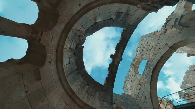 Smooth rotating camera movement looking upward inside Alahan Monastery in Mut, Mersin, T&uuml;rkiye, revealing ancient stone arches, columns, weathered walls and open blue sky. Historic early Christian arc