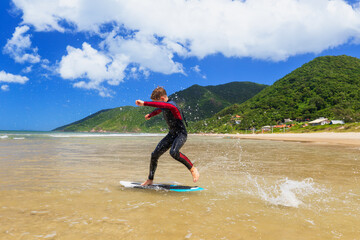 Fototapeta premium Young boy skimboarding in shallow water on a sunny beach day. Wearing wetsuit, balancing on board with determination and focus. Perfect image for themes of childhood adventure, active lifestyle