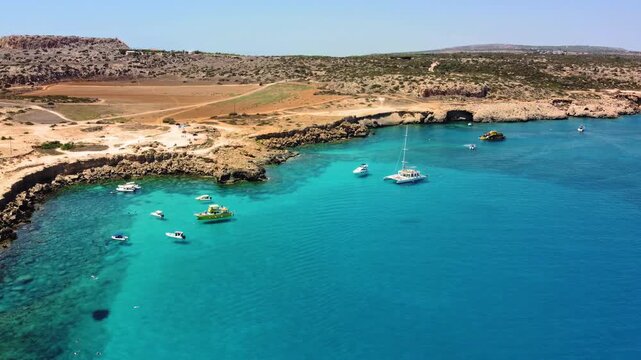 Aerial view of boats anchored in crystal clear turquoise water near a rugged, sun-drenched coastline with sparse vegetation under a clear blue sky. Aerial coastal landscape.