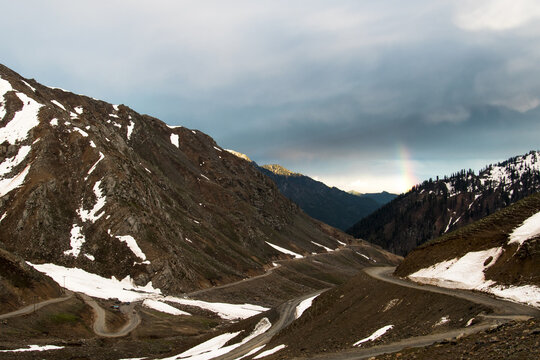 View of the winding road at Lowari Pass surrounded by snow-capped mountains and rugged terrain under a dramatic sky with a rainbow in Khyber Pakhtunkhwa, Pakistan.