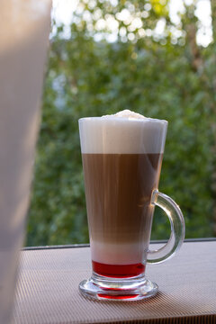 Tall glass of coffee with raspberry syrup and milk foam on table with curtain in foreground. Cozy window morning beverage scene.