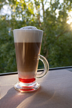 Tall glass mug with layered coffee with raspberry syrup, milk, espresso, and thick milk foam on a table by a window in sunlight. Fresh morning drink concept.