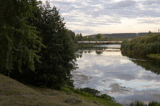 View along the grassy bank of the Ukhta River towards an industrial pipeline bridge in Ukhta city, Komi Republic, Russia. Calm water reflecting sky.
