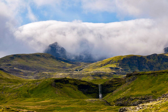 View of Eyjafjallajokull volcano partially covered by clouds with lush green slopes and a small waterfall in the foreground in Eyjafjallajokull, Rangarthing eystra, Iceland.