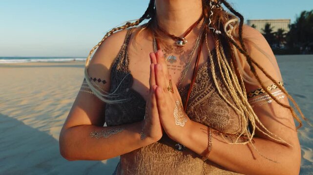Woman with dreadlocks and temporary tattoos meditating on a beach, finding calm and spiritual connection with hands in a prayer pose against a backdrop of ocean waves and golden sand