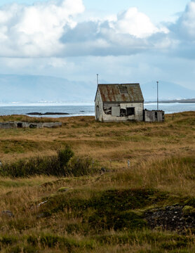 View of an abandoned house with a rusty roof set in a grassy field near the coast with mountains in the distance in Eyjafjallajokull, Rangarthing eystra, Iceland.