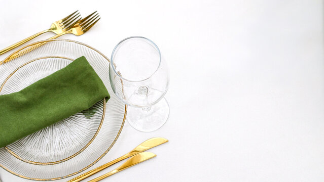 Elegant table setting with clear textured glass plates, a folded green linen napkin, shiny gold cutlery, and an empty wine glass on a clean white tablecloth, offering copy space